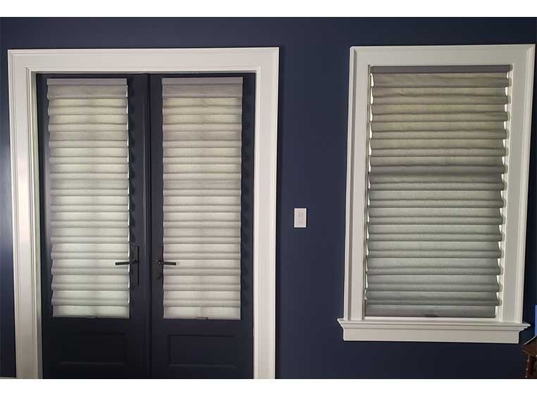 A double glass door next to a tall window, both covered  by roman shades